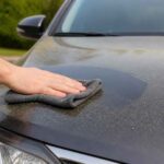 Dłoń przeciera zakurzoną maskę samochodu szarą ściereczką microfiber.