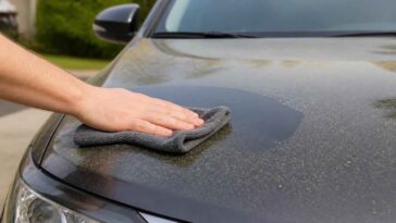 Dłoń przeciera zakurzoną maskę samochodu szarą ściereczką microfiber.